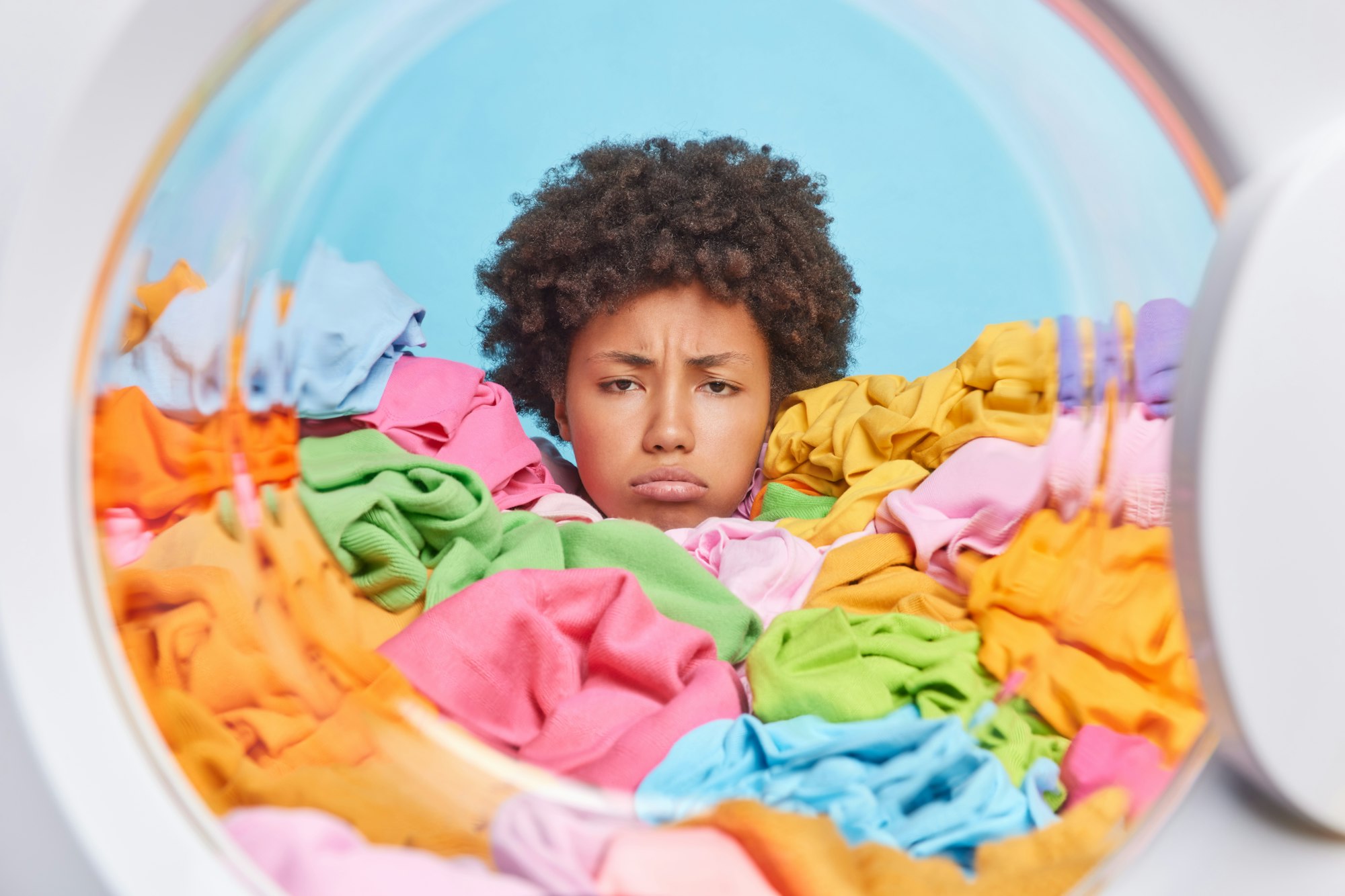 Unhappy African American woman with curly hair has tired expression does laundry at home drowned in