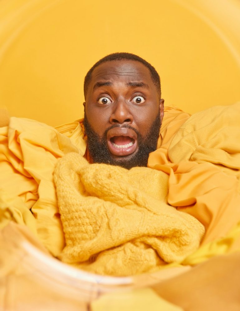 Shocked bearded African American man busy houseworking stares bugged eyes drowned in dirty laundry p